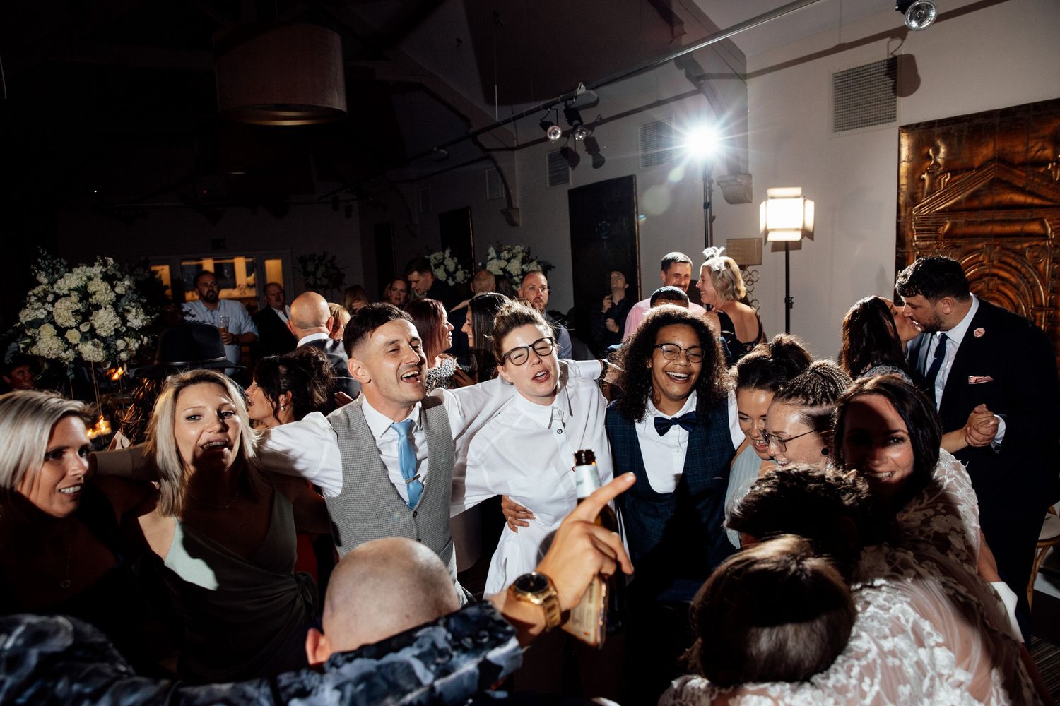 Wedding reception guests dance and celebrate on a dark dance floor with dramatic lighting.