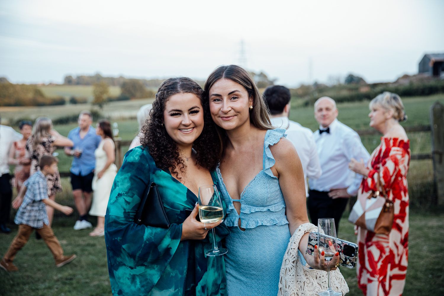 Two guests in elegant dresses enjoying an outdoor wedding celebration with other attendees in the background.