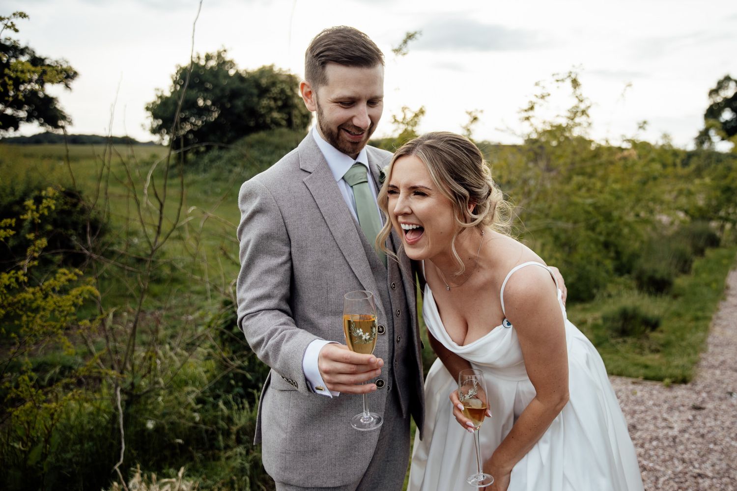 A couple shares a joyful laugh while holding drinks at their outdoor wedding celebration.