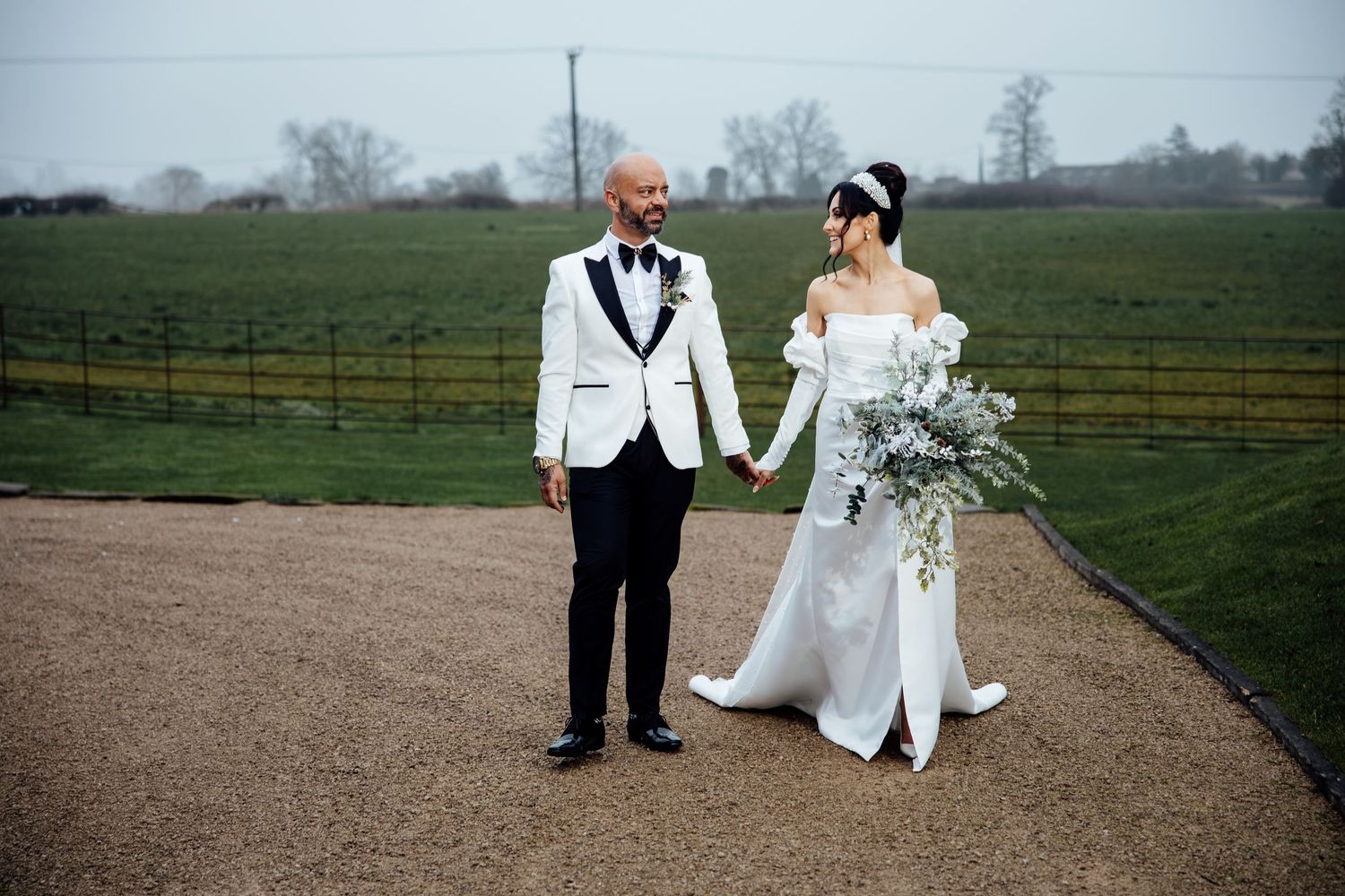 Bride and groom in white formal attire pose on a path with countryside views.