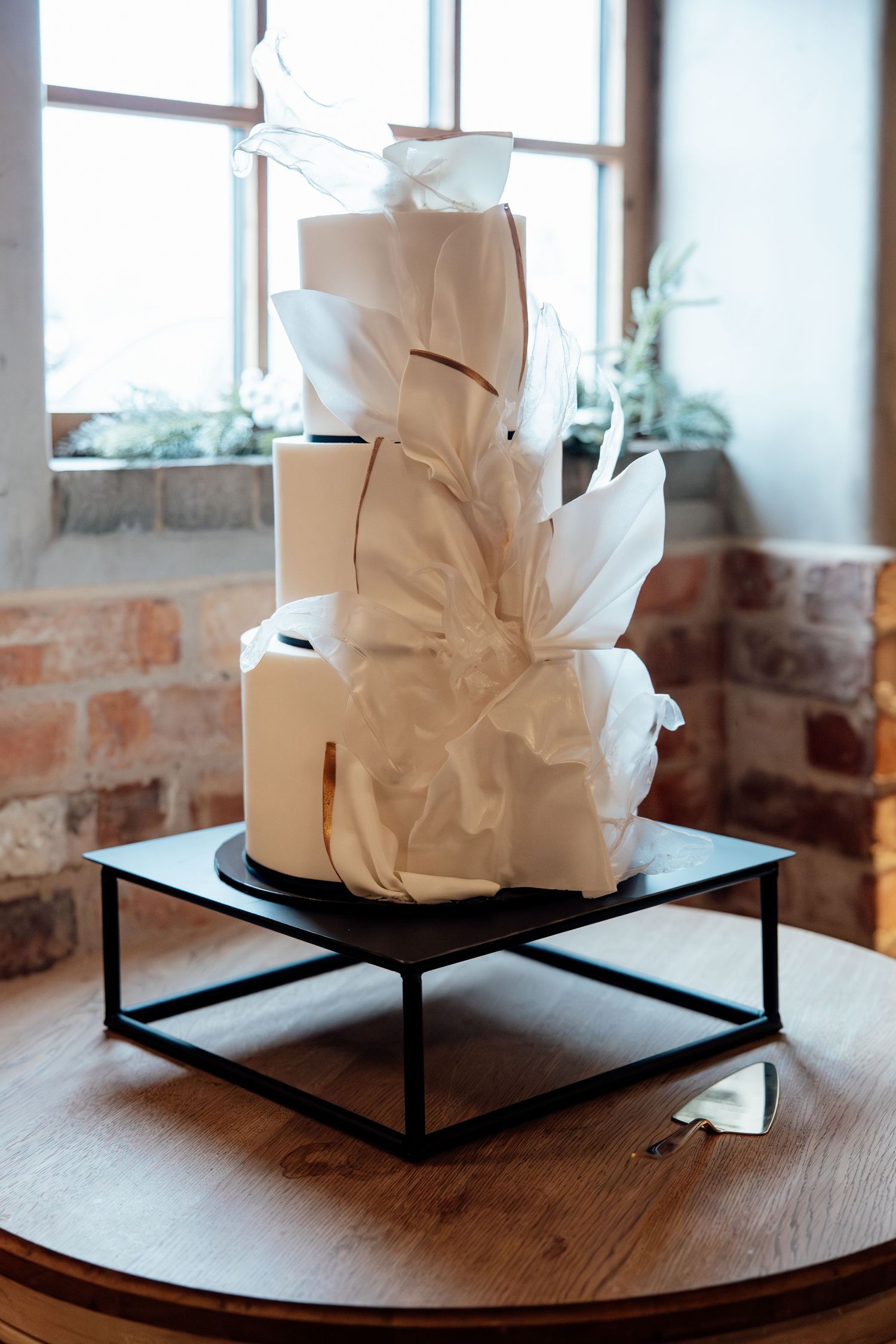 Modern white wedding cake with abstract geometric paper decorations displayed on black metal stand.