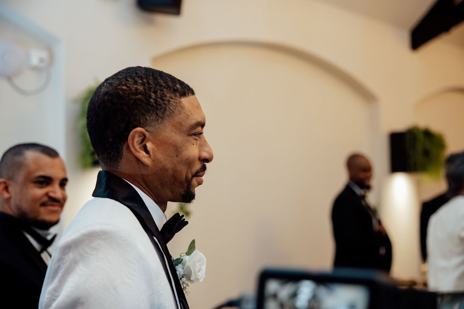 Groom in white tuxedo with boutonniere smiling during wedding ceremony.