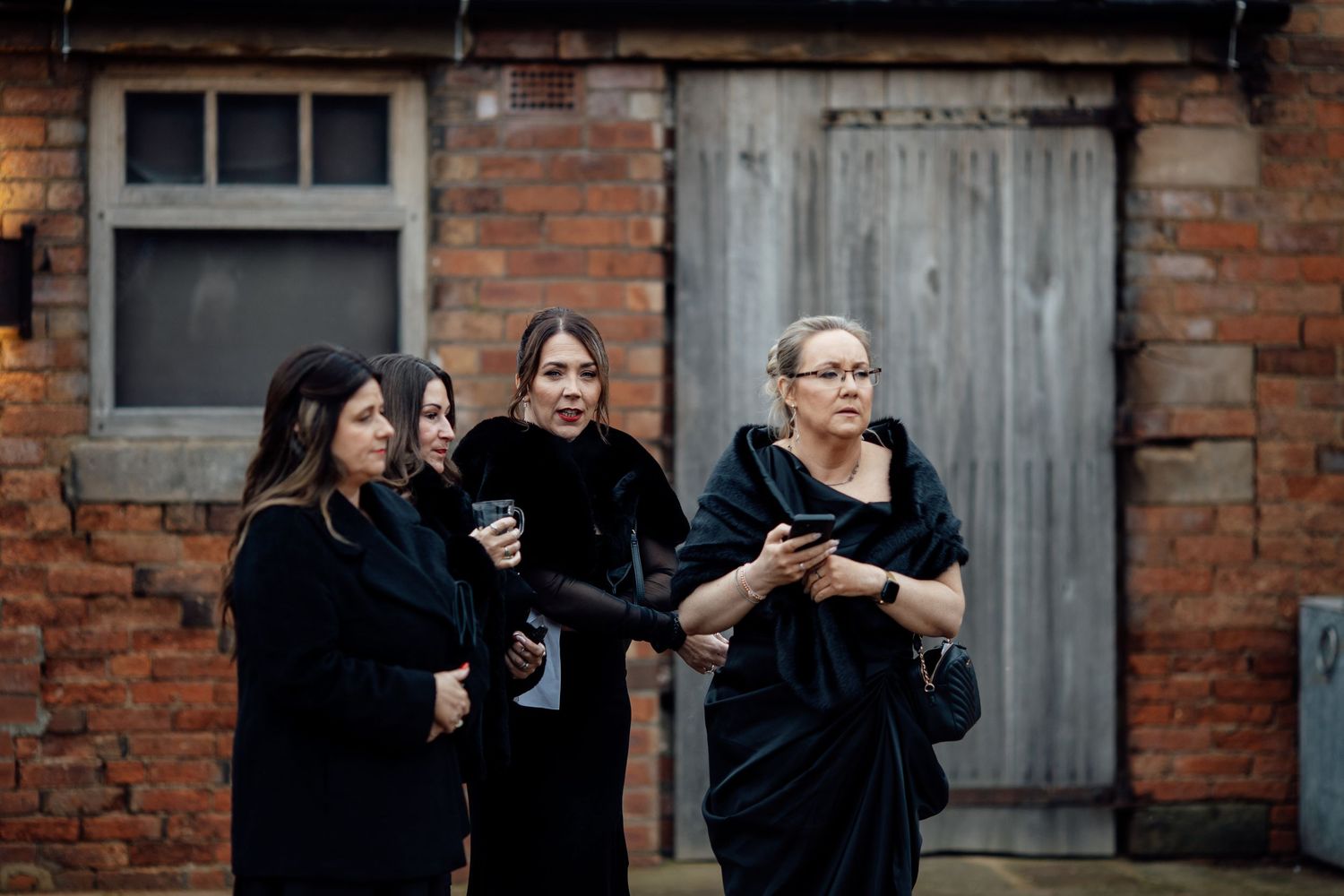 Three wedding guests in dark formal attire checking their phones outside a brick building.