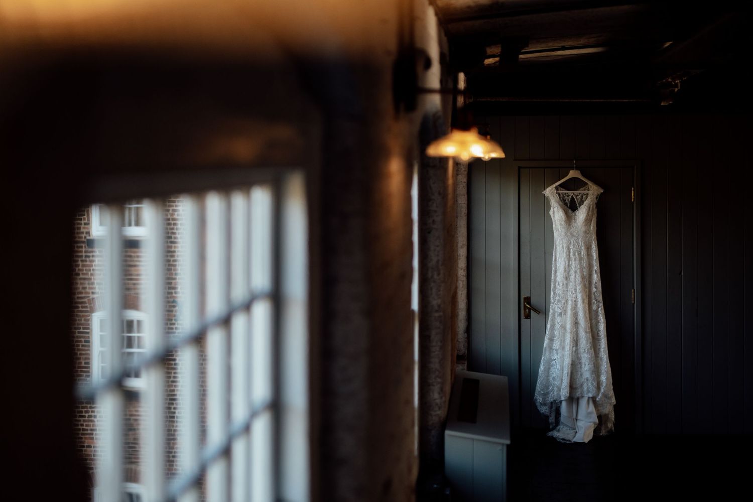 Wedding dress hangs elegantly by a window in natural light during bridal preparations.