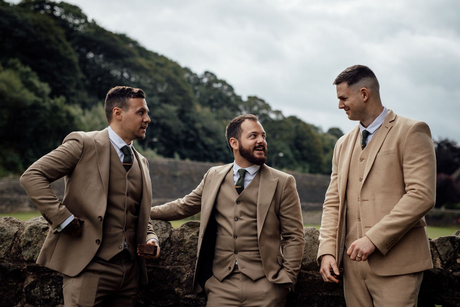 Groomsmen in tan suits pose together outdoors against scenic hillside backdrop.