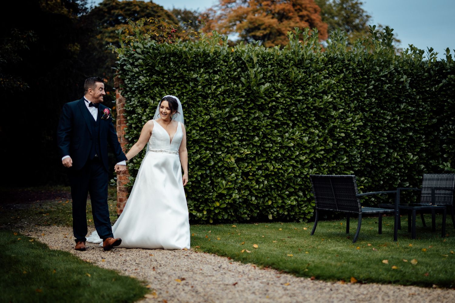 Couple in wedding attire walking along a scenic garden path during autumn.