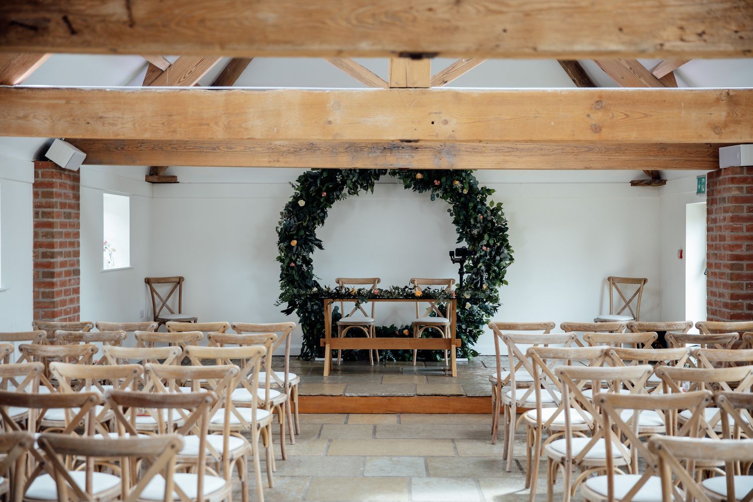 A minimalist wedding ceremony setup with greenery arch and cross-back chairs in a bright venue.
