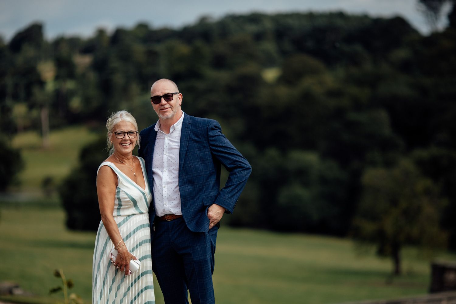 Couple poses in formal attire against rolling countryside landscape.