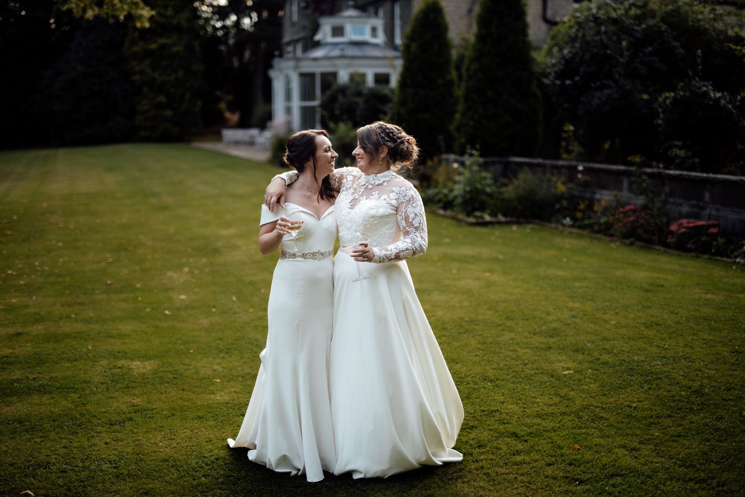 Two people in white dresses walking together on a manicured lawn with trees in the background.