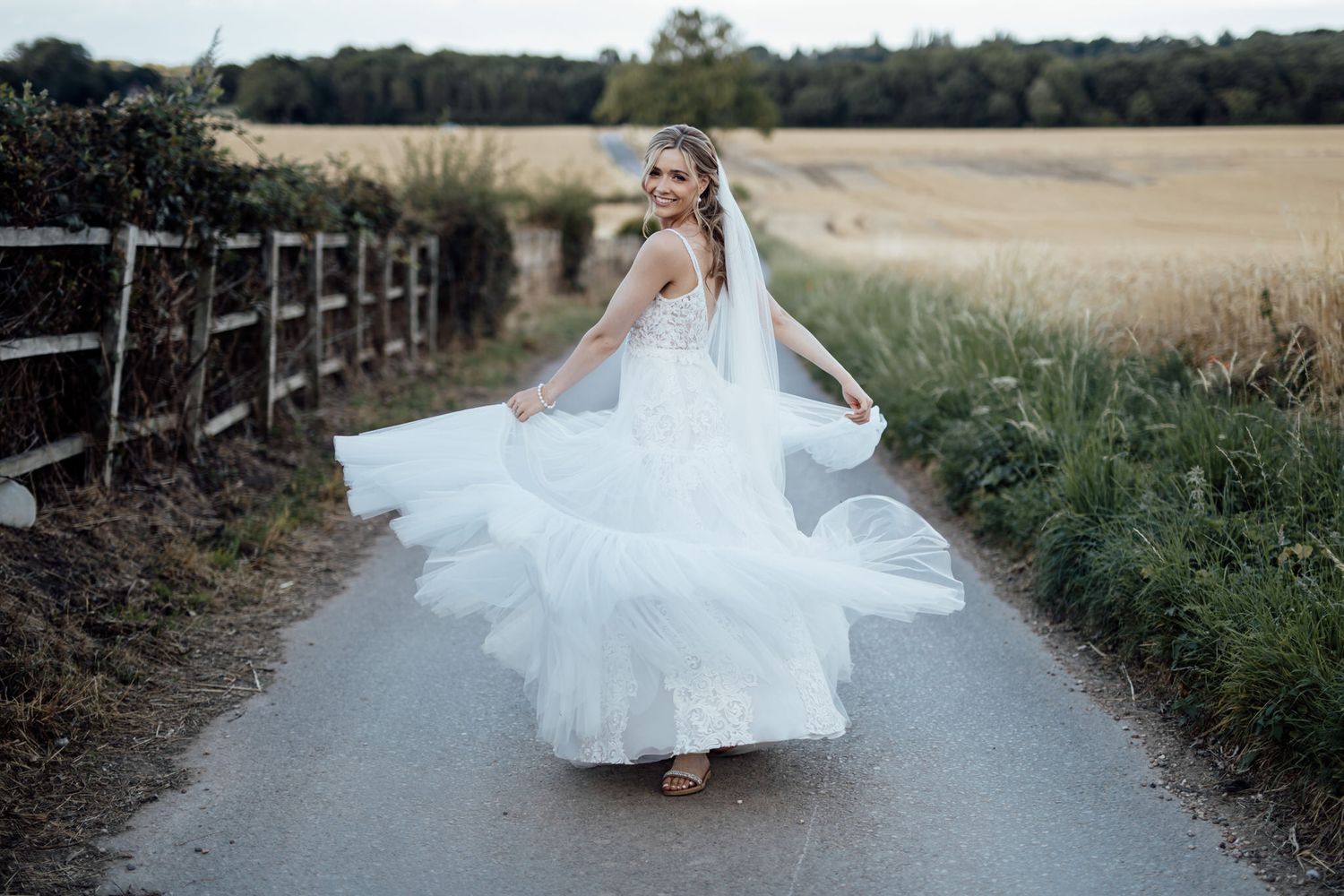 Bride twirls white wedding dress on country path bordered by wooden fence and wheat field.
