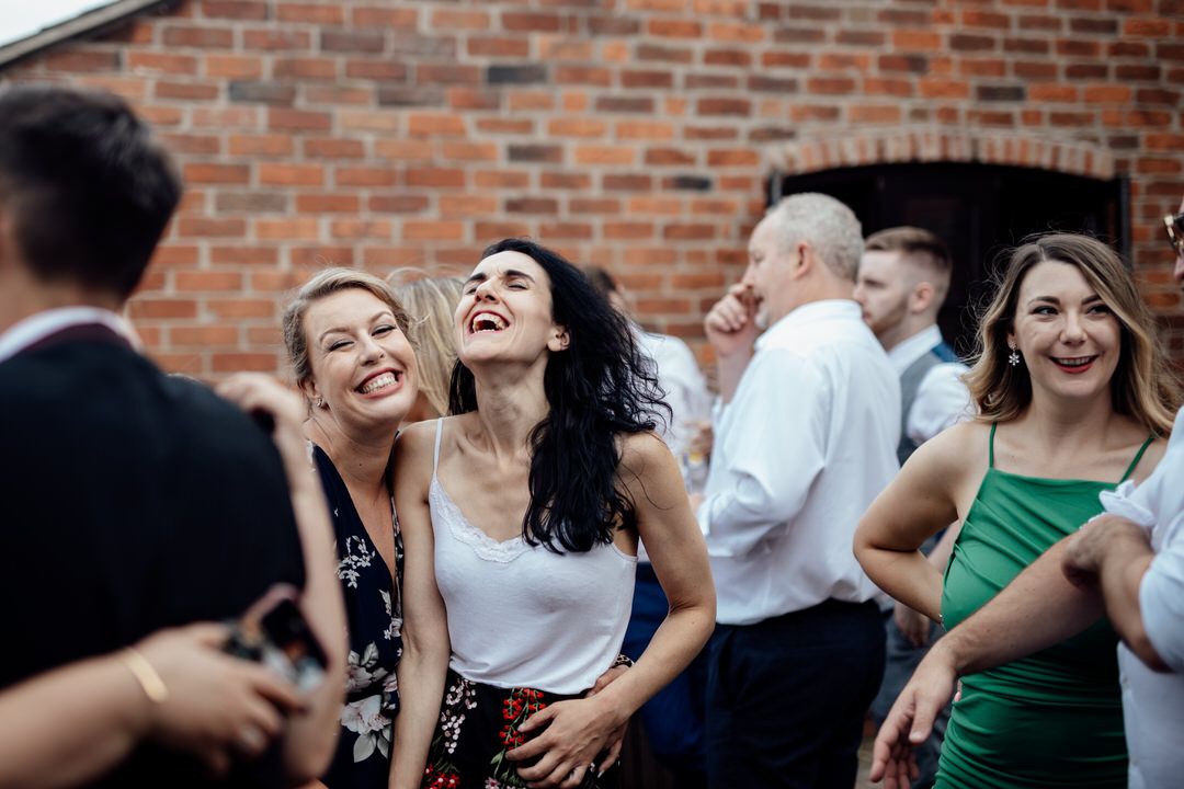 Wedding reception guests laugh and dance together against exposed brick wall backdrop.