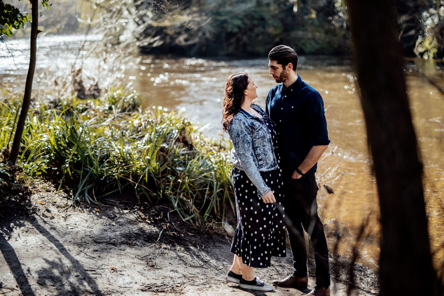 Two people share a tender moment by the water's edge in a serene park setting.