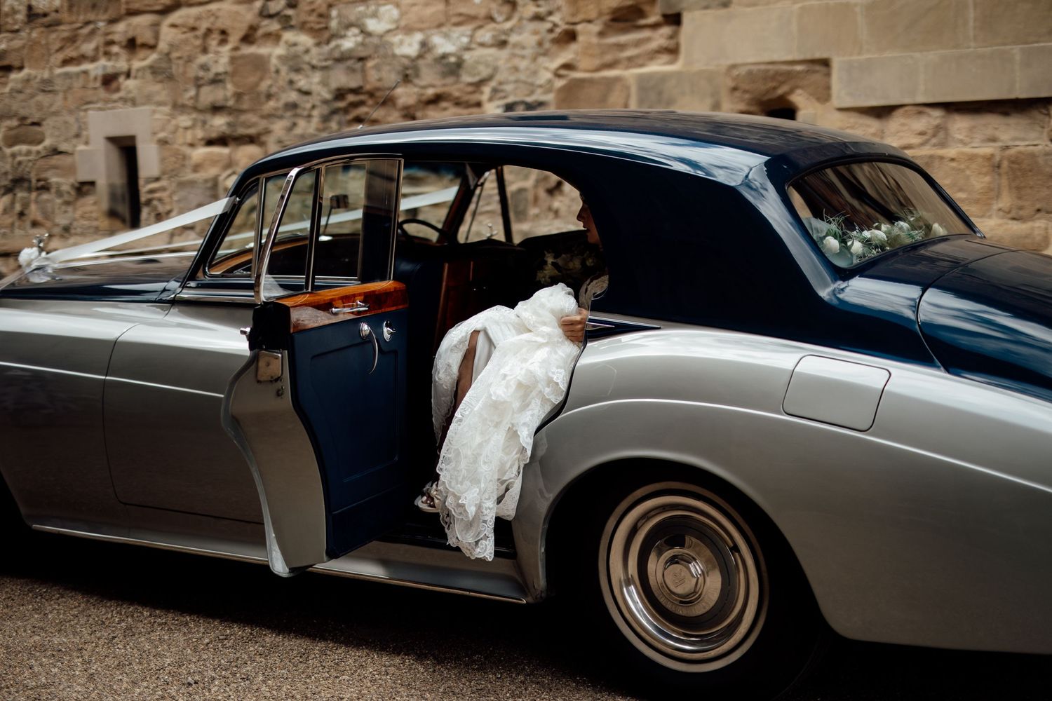 Classic vintage wedding car in silver with white details parked outside stone building.