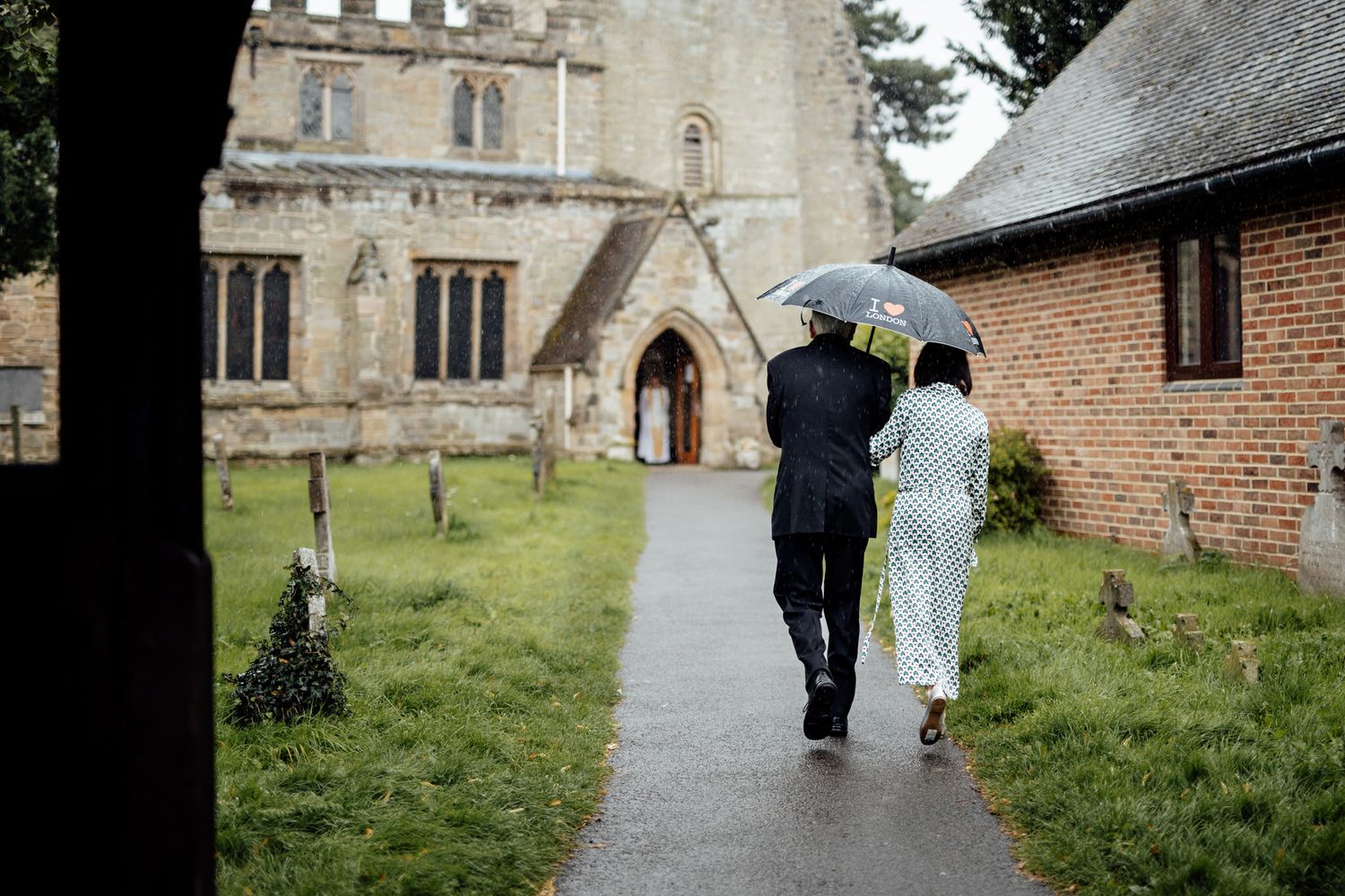 A couple walks through a rainy church courtyard holding umbrellas on their wedding day.