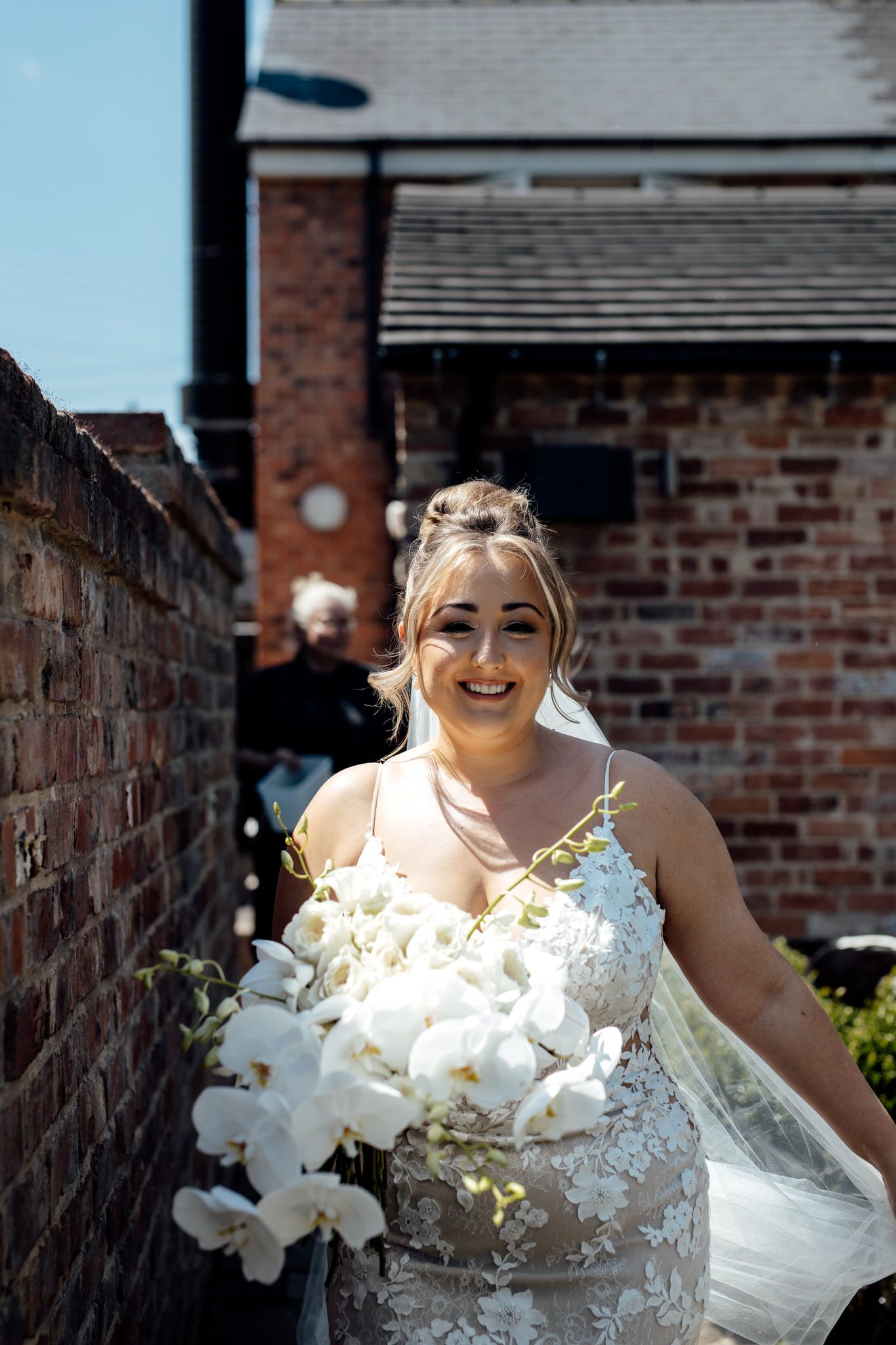 Bride in lace dress with white orchid bouquet poses against brick wall.