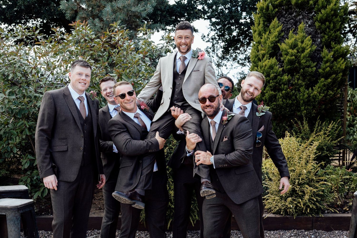 Groomsmen in dark suits lift the groom outdoors in a fun wedding celebration photo.