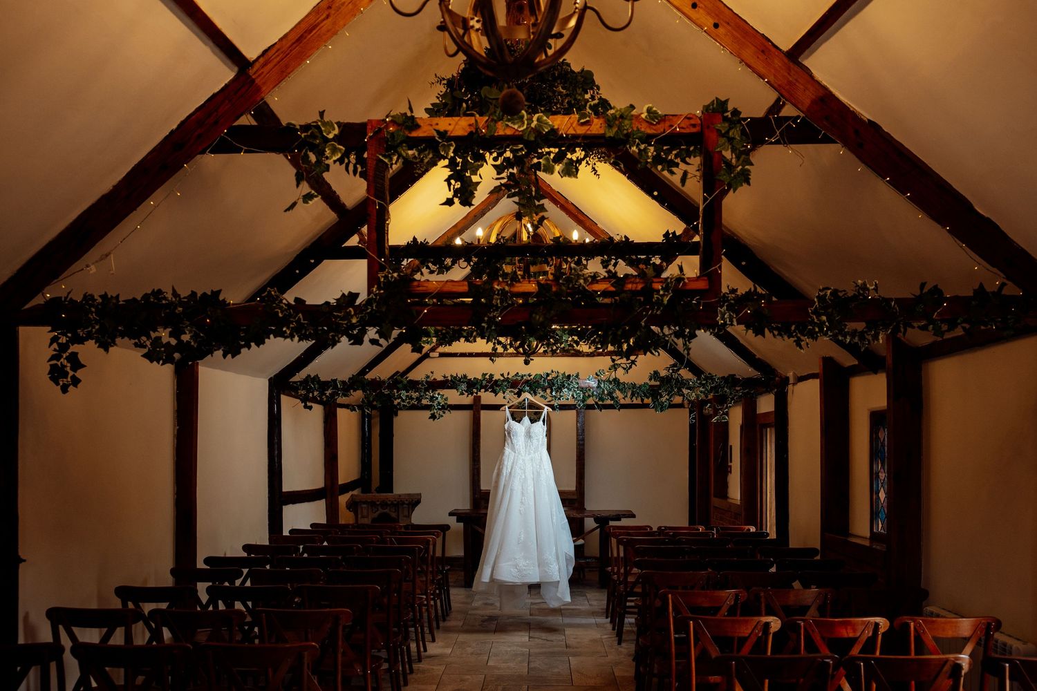 Wedding dress hanging in a rustic barn chapel adorned with greenery garlands and warm lighting, with wooden chairs arranged in rows.