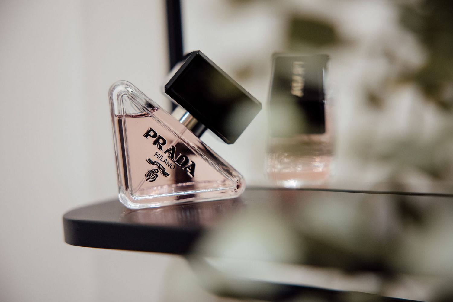 Prada Paradoxe perfume bottle in pink triangular glass on a dark shelf with blurred greenery.