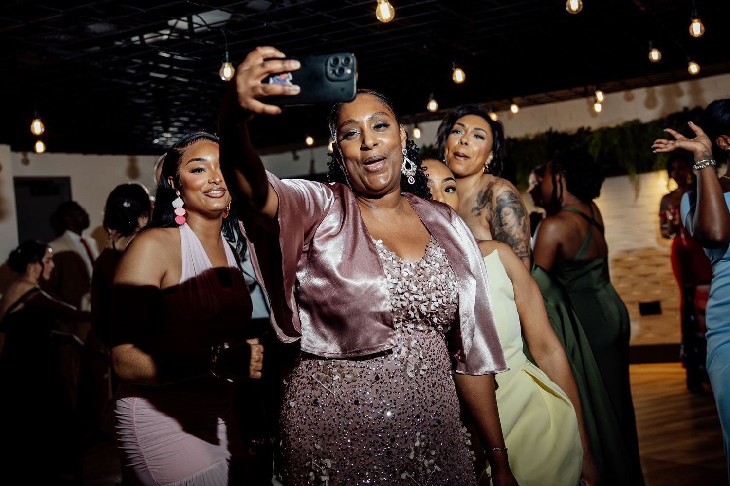 Women in elegant dresses taking a selfie while dancing at a festive indoor event with string lights.