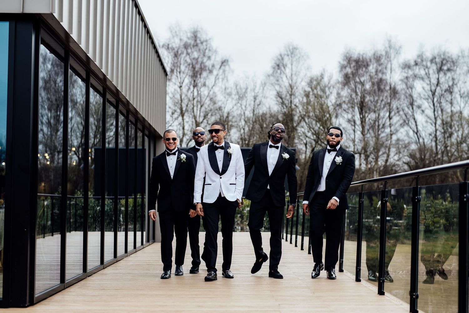 Groom in white tuxedo poses with groomsmen in black suits on modern glass building deck.