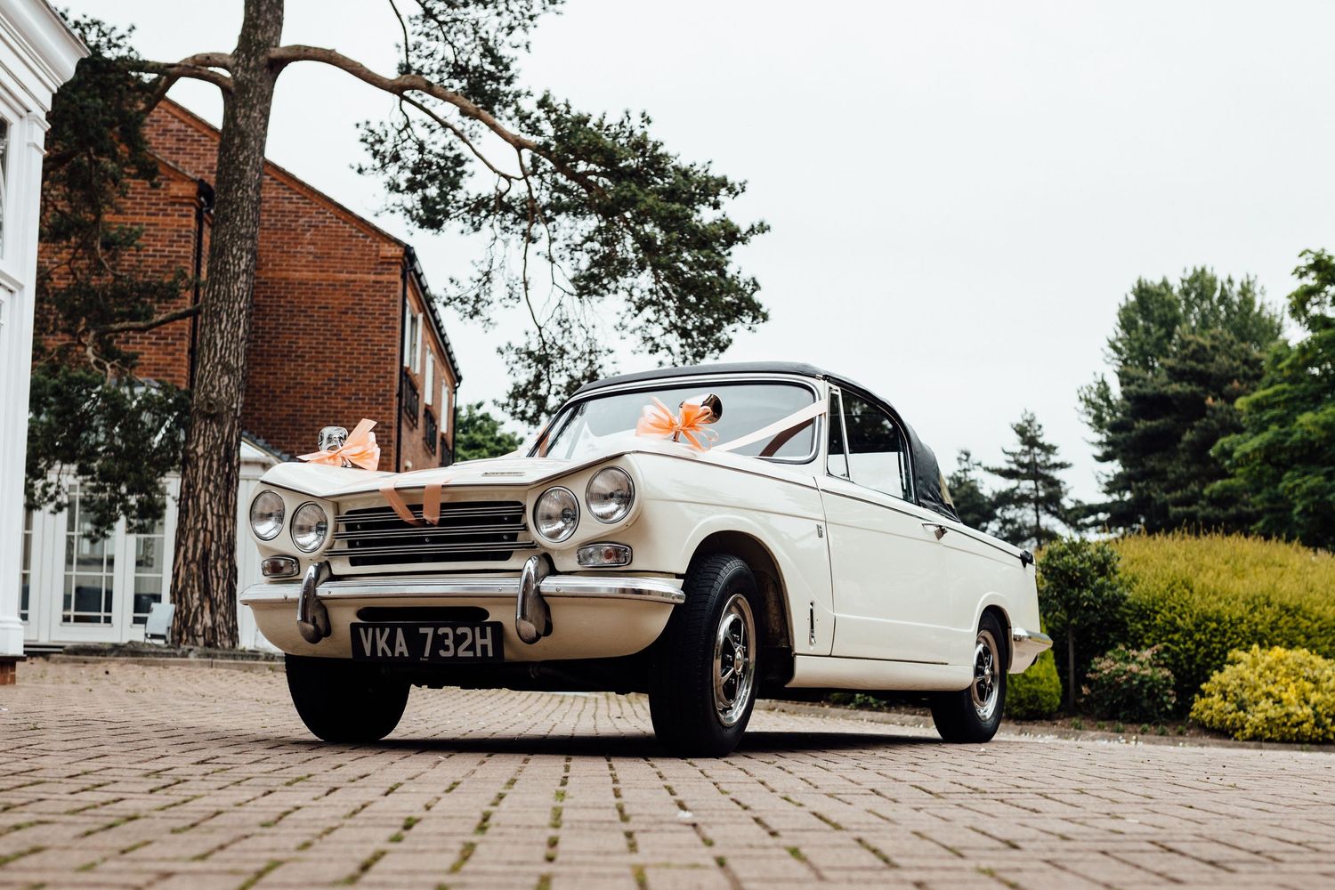 Vintage white wedding car parked on brick pavers near garden landscaping.