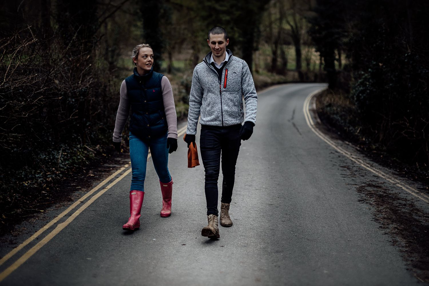 A couple walking down a paved country road surrounded by trees.
