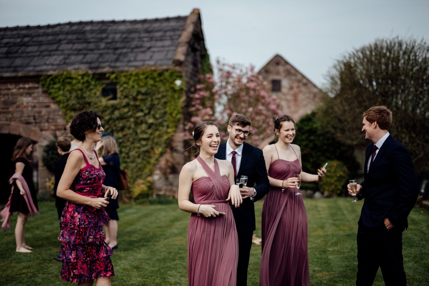 Wedding party in burgundy dresses and dark suits poses on lawn near brick building.