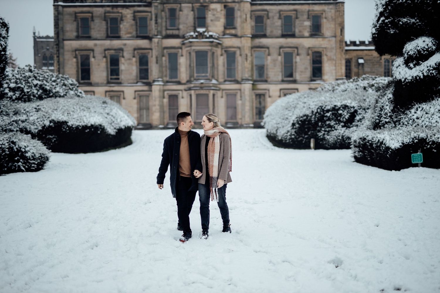 A couple walks through snow in front of a grand historic mansion with snow-covered gardens.