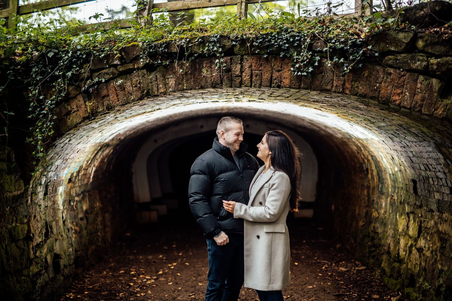 Two people stand close together under a rustic stone bridge tunnel surrounded by greenery.