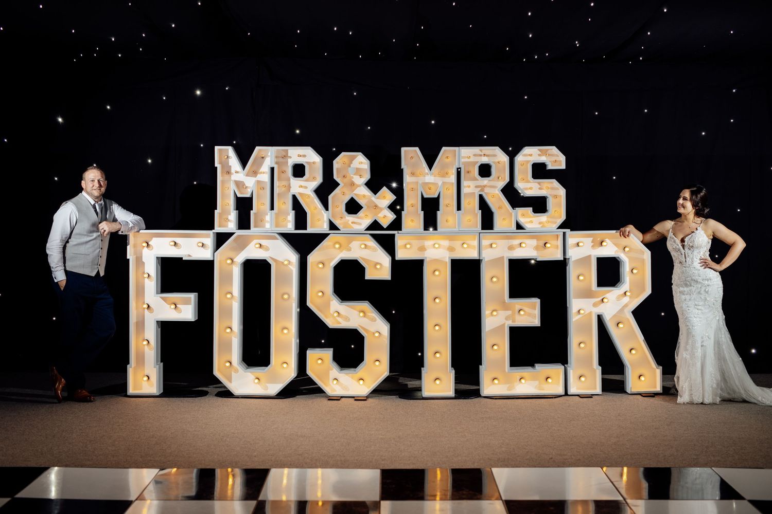 Large illuminated 'MR & MRS FOSTER' sign displayed at a wedding reception against a starry backdrop.