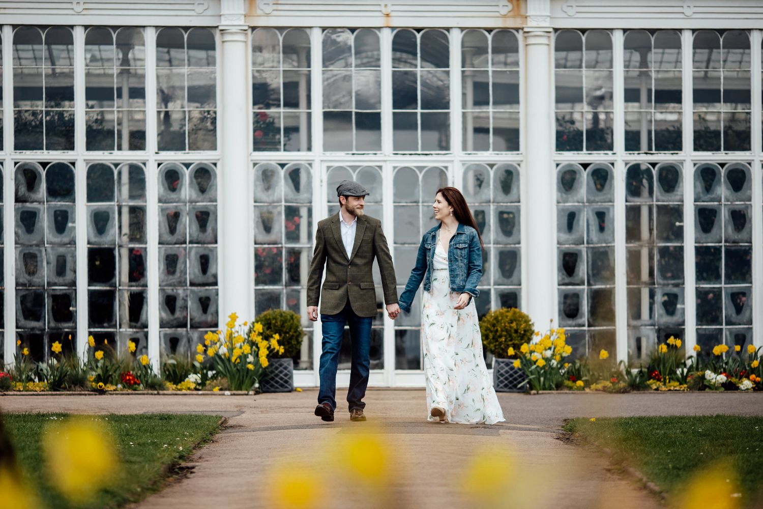 A couple walks along a path in front of a grand conservatory building with glass windows and yellow flowers.