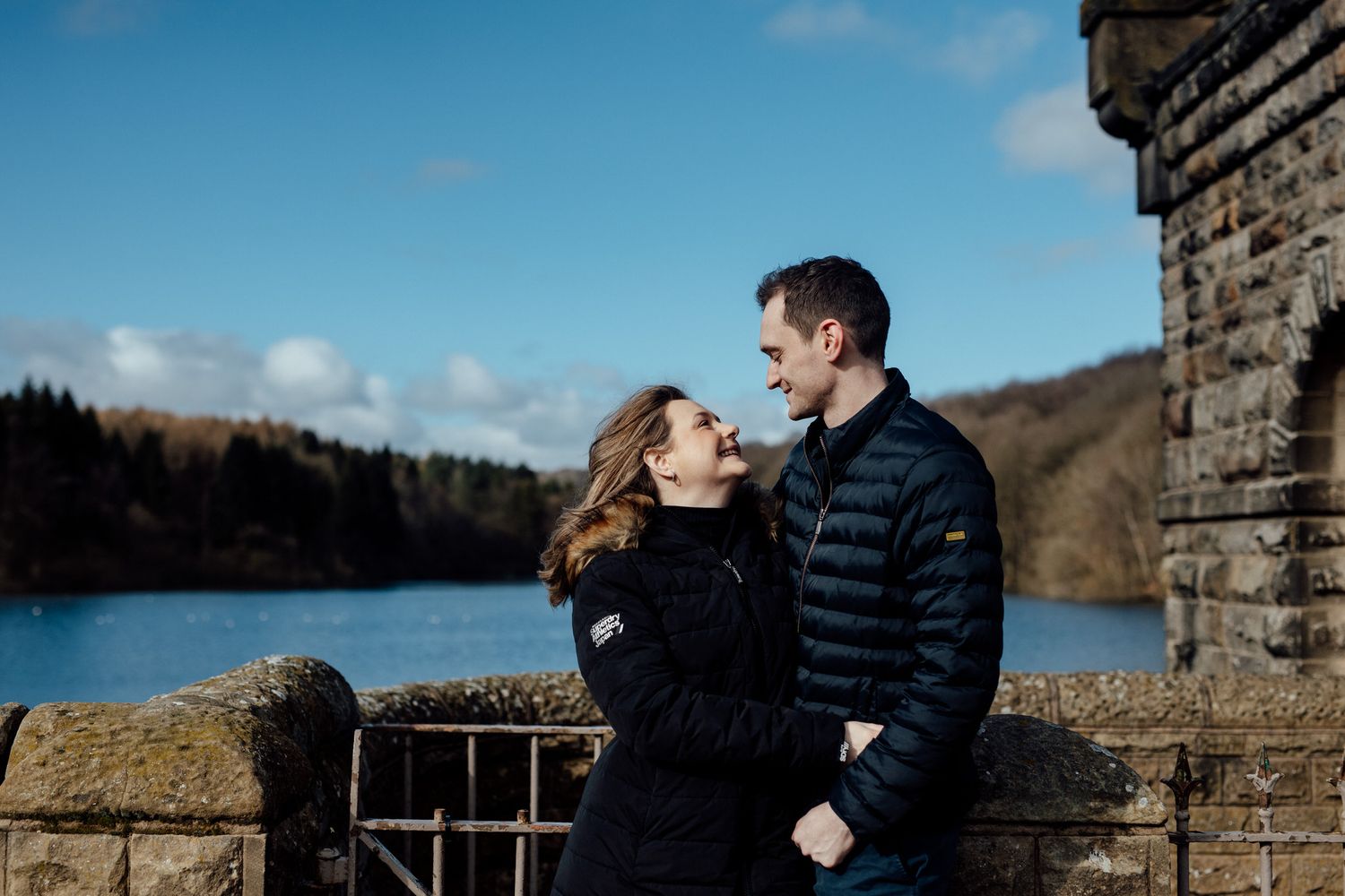 A couple walks together along a stone wall path on a sunny winter afternoon in a scenic outdoor setting.