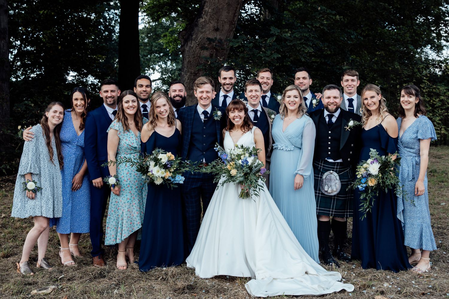 Wedding party group photo showing bridesmaids in light blue dresses and groomsmen in navy suits outdoors.