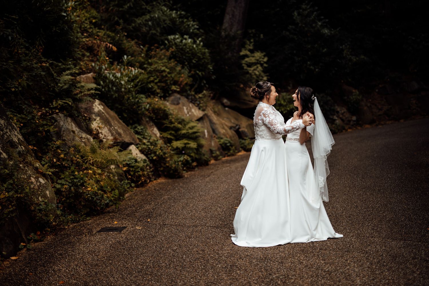 Two brides in white wedding dresses walking together on a forest path.
