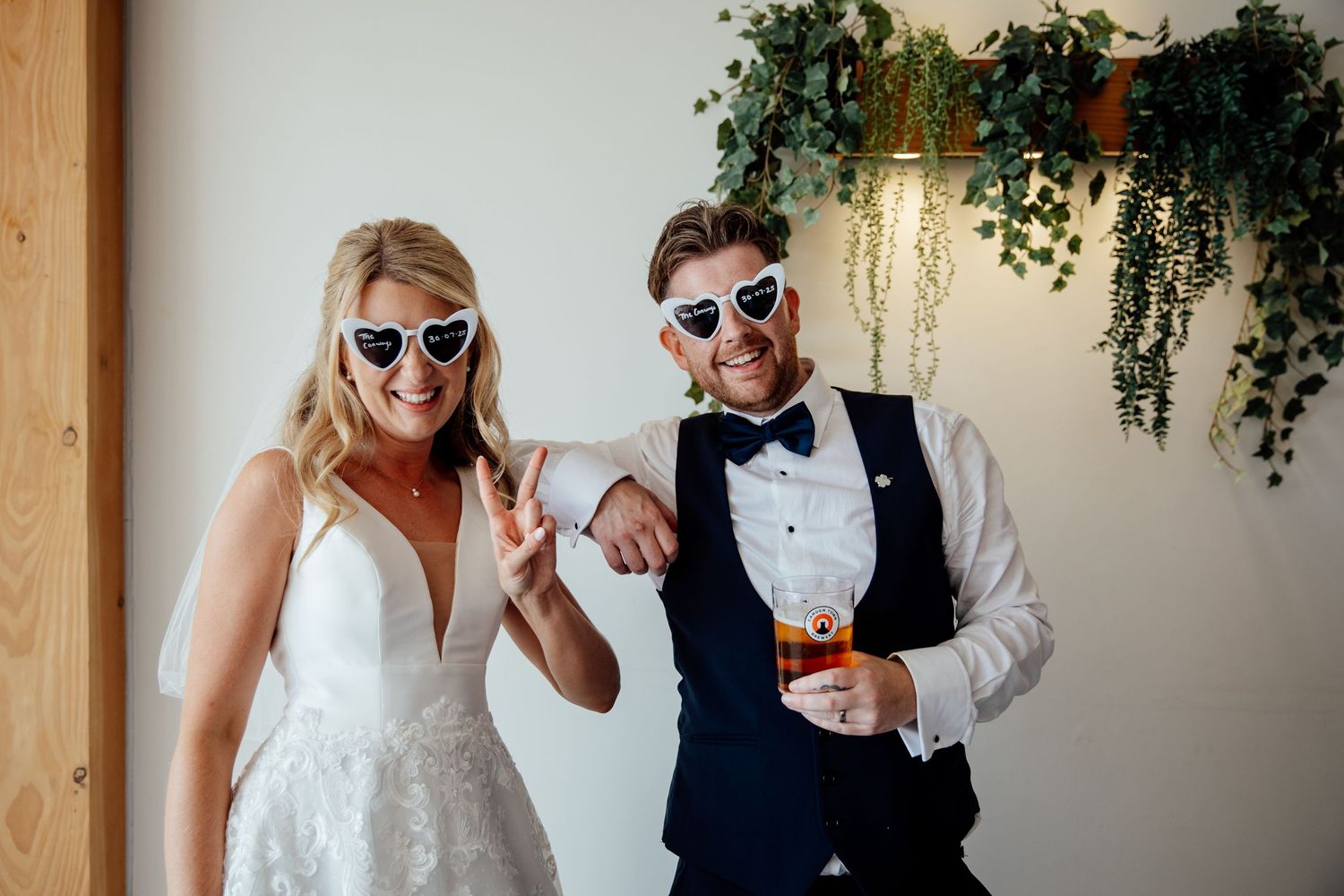 Wedding couple poses playfully with sunglasses indoors under hanging greenery.