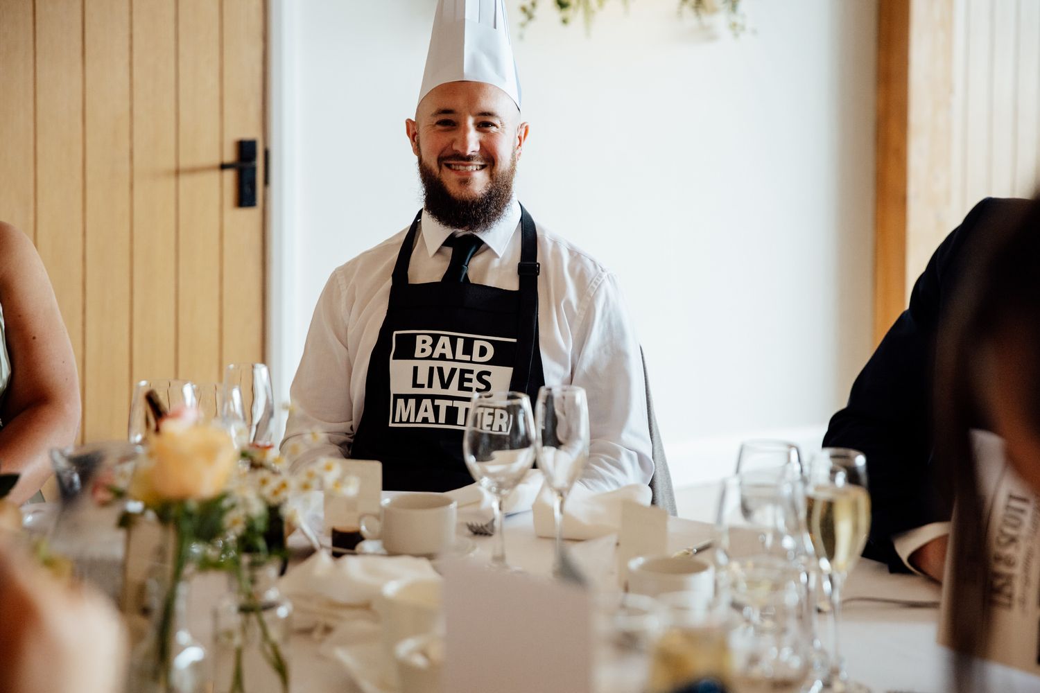 Server wearing 'Bake Love Not Hate' apron stands at wedding reception table.
