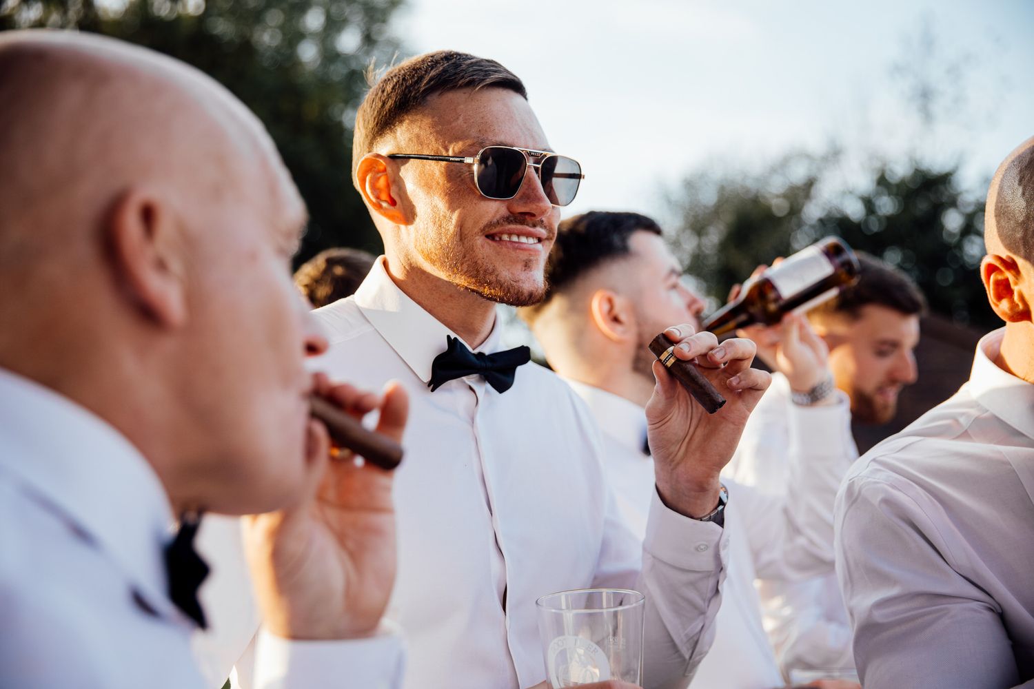 Groomsmen in bowties and sunglasses enjoying drinks during outdoor wedding celebration.