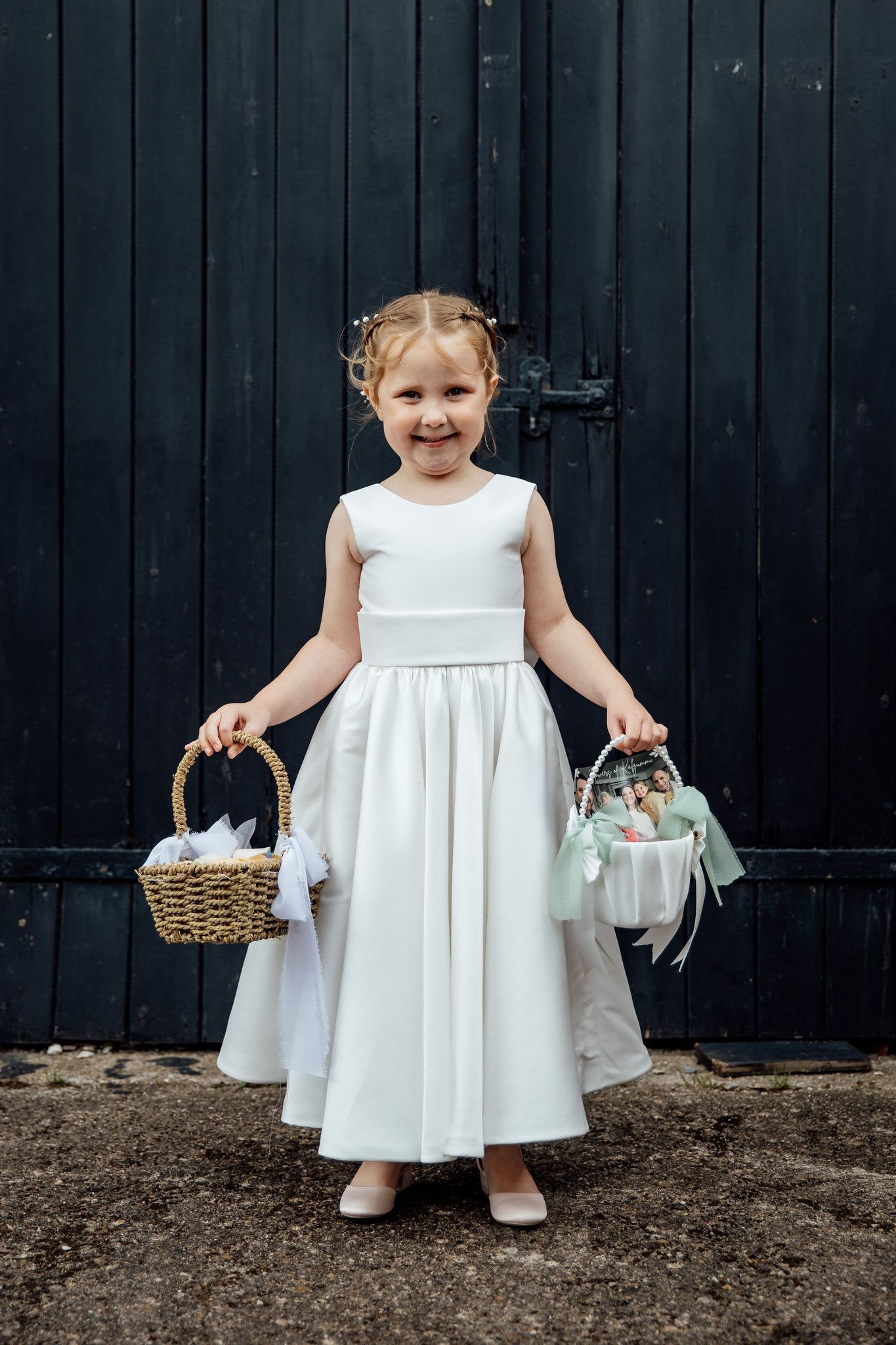 A flower girl in a white dress holds two decorative baskets against a dark wooden door.