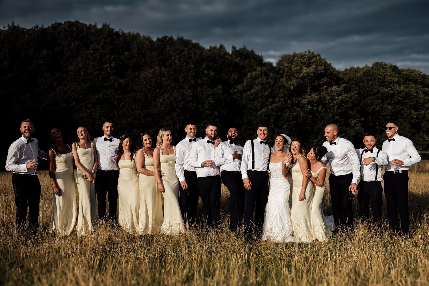 Wedding party in cream dresses and white shirts poses in dramatic formation against moody sky.