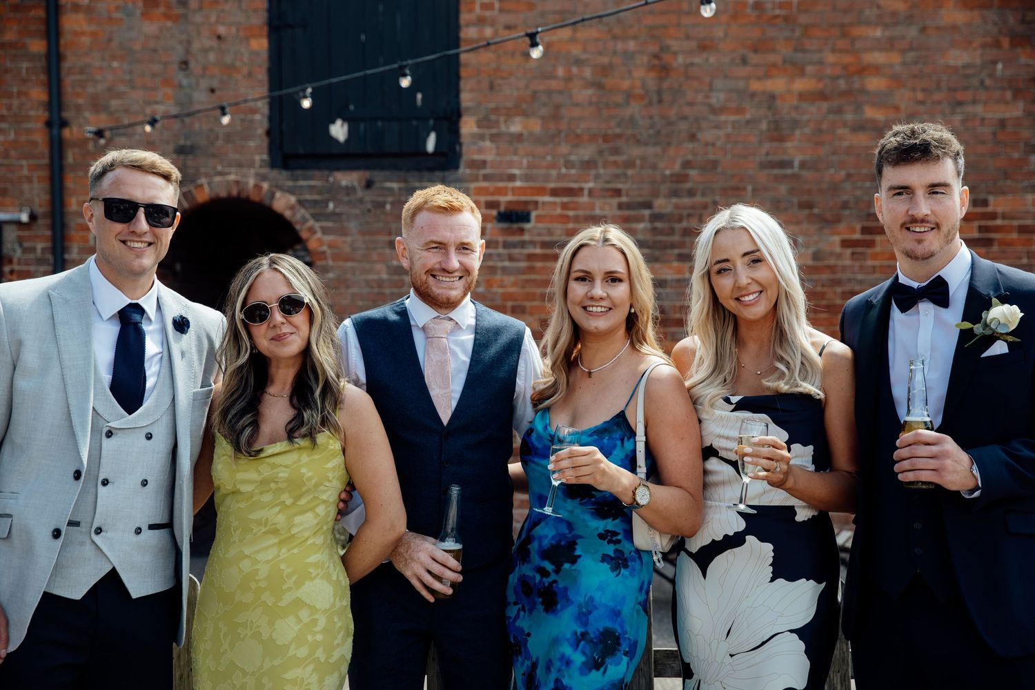 Wedding party posing together outside brick building with string lights.
