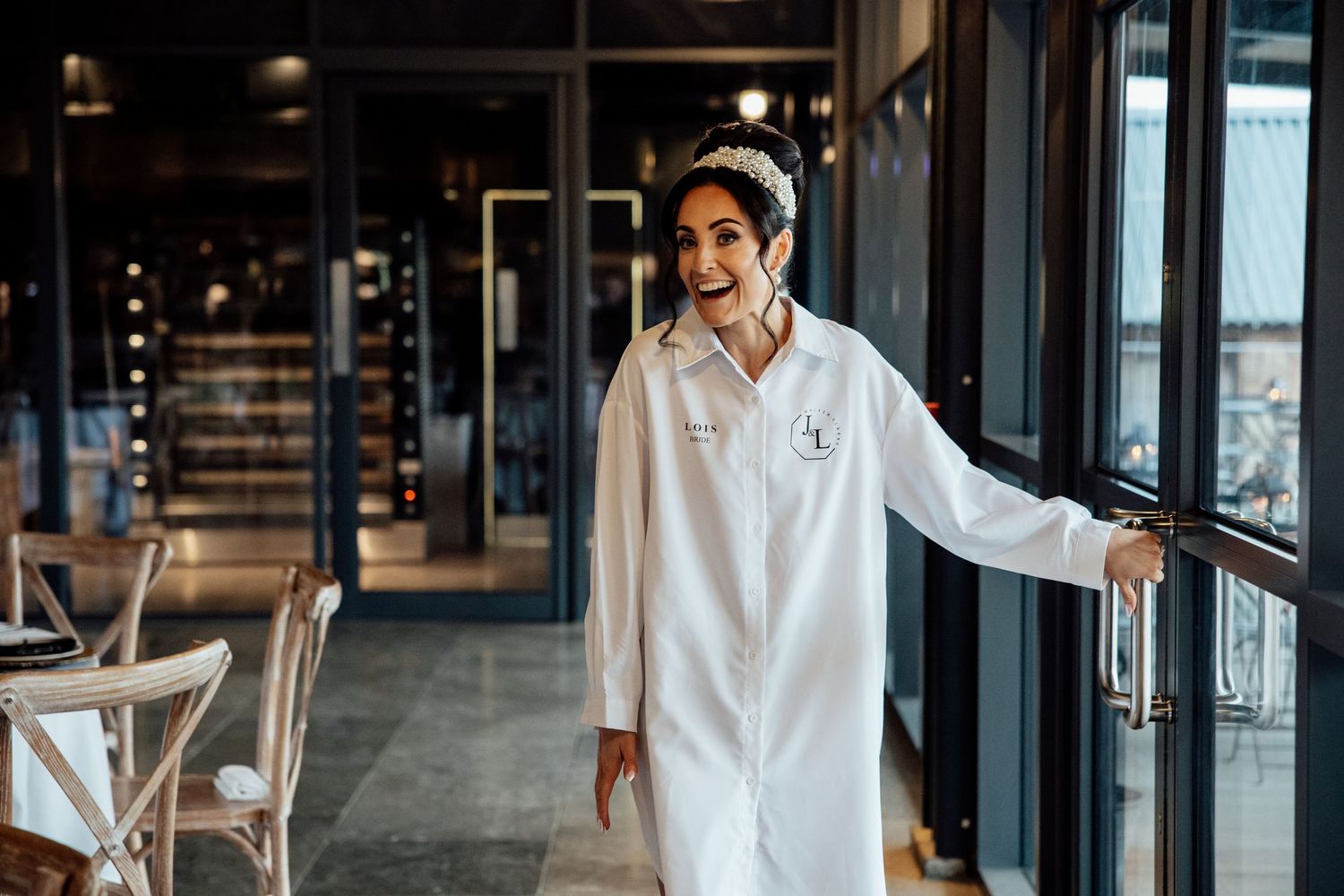 A chef in white uniform stands smiling in a modern restaurant interior with large windows.