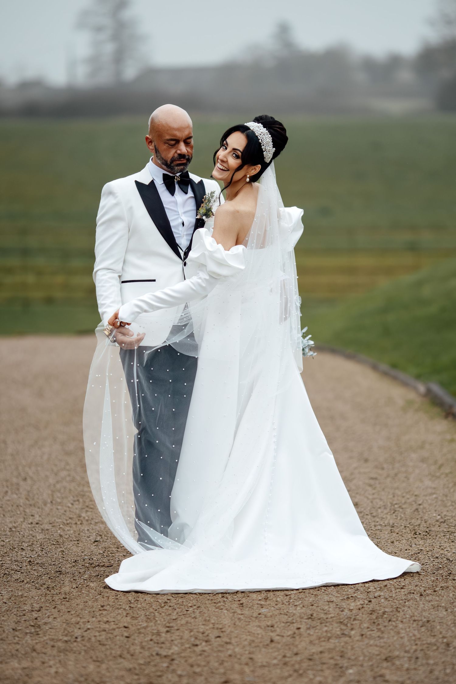 Newlyweds pose for romantic portraits on a dirt path with the bride's veil flowing dramatically in the wind.