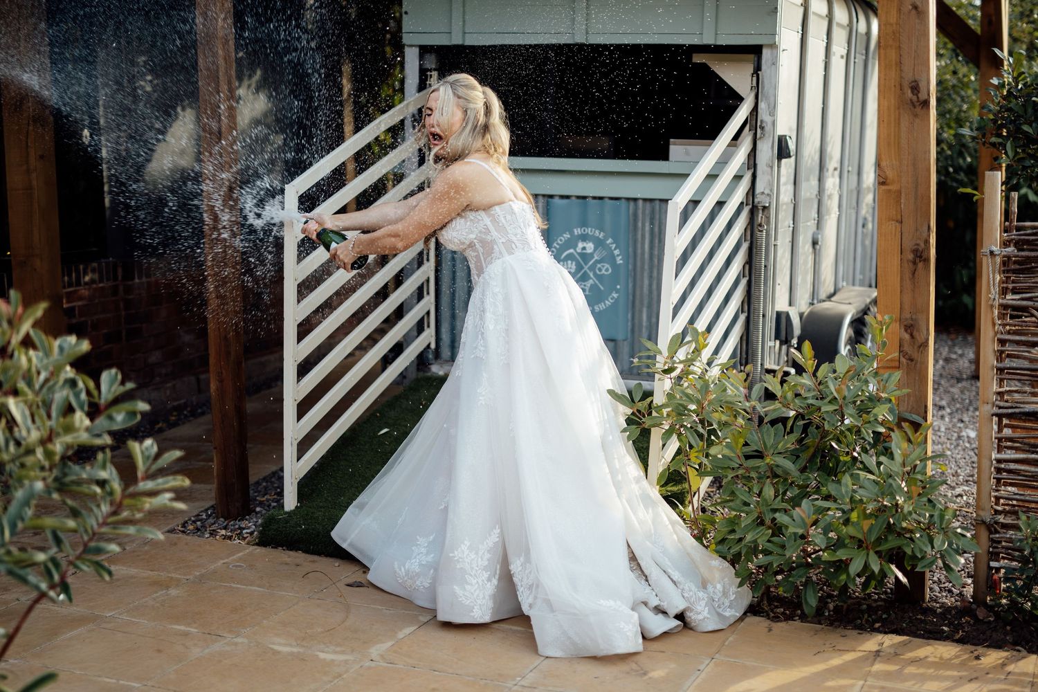 A bride in a flowing white ball gown tosses confetti outdoors near a garden gate.