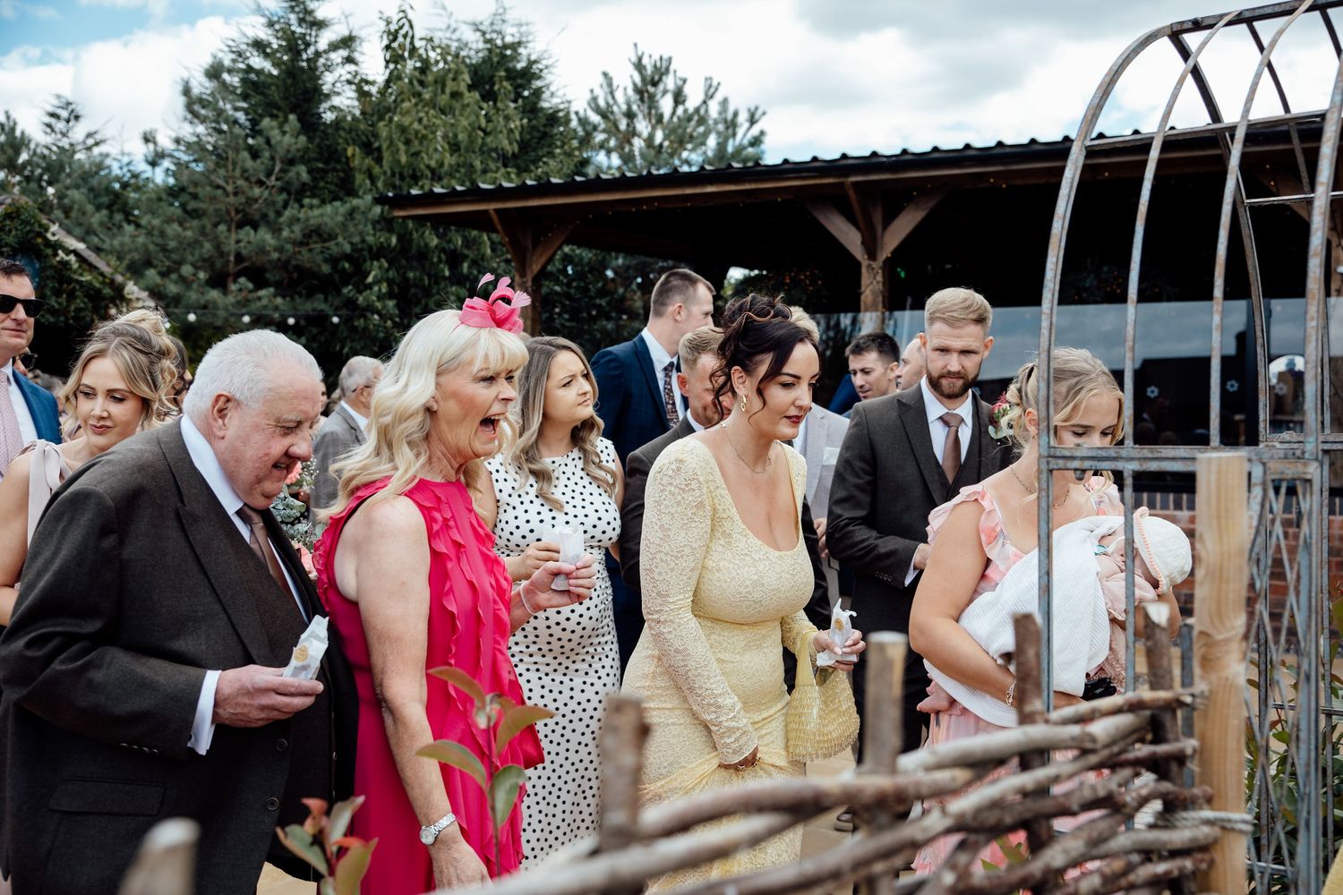 Wedding guests mingle outdoors at a rustic venue, dressed in elegant attire including a bright pink dress.