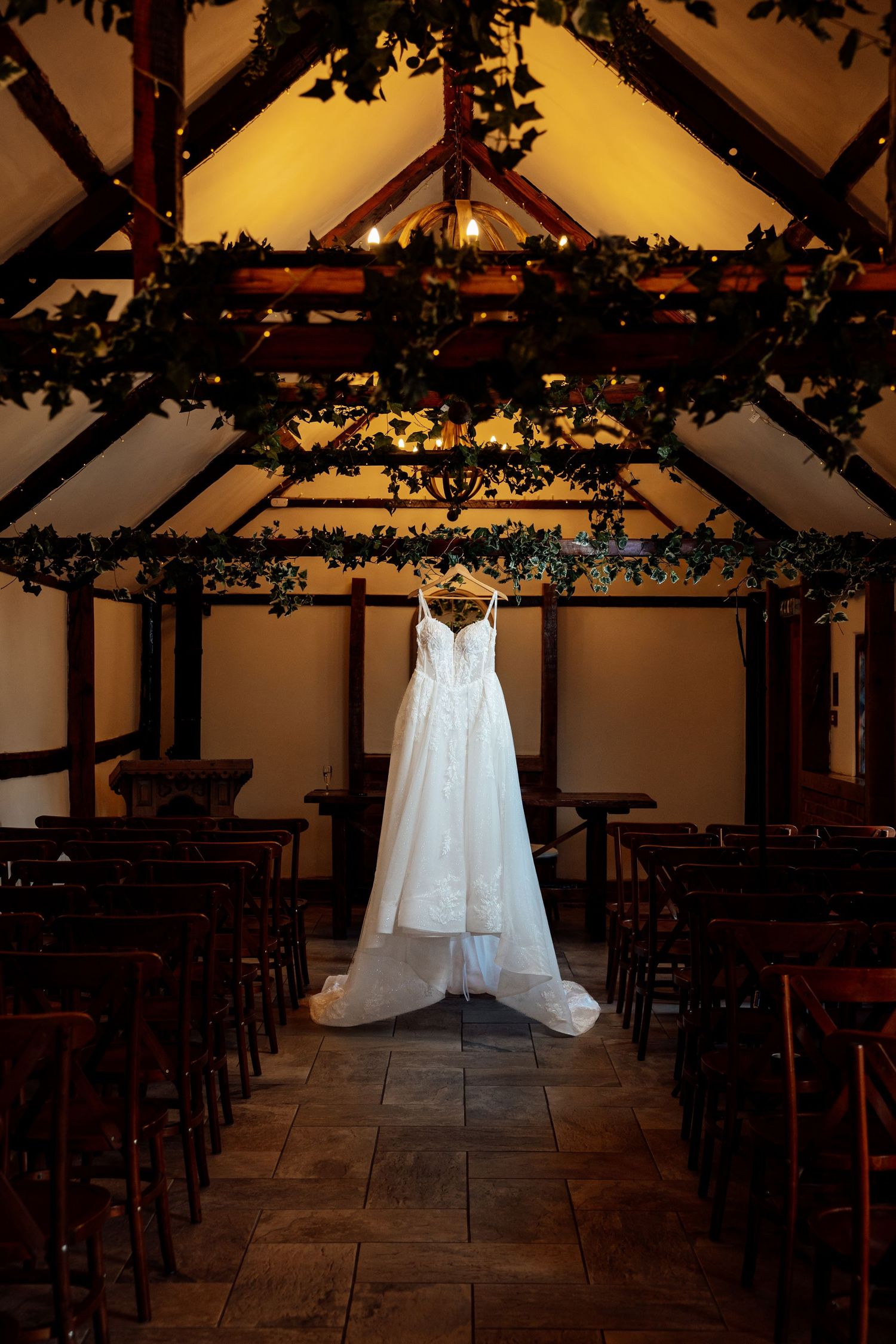 Elegant white wedding dress hanging in a rustic barn venue with green foliage decor.
