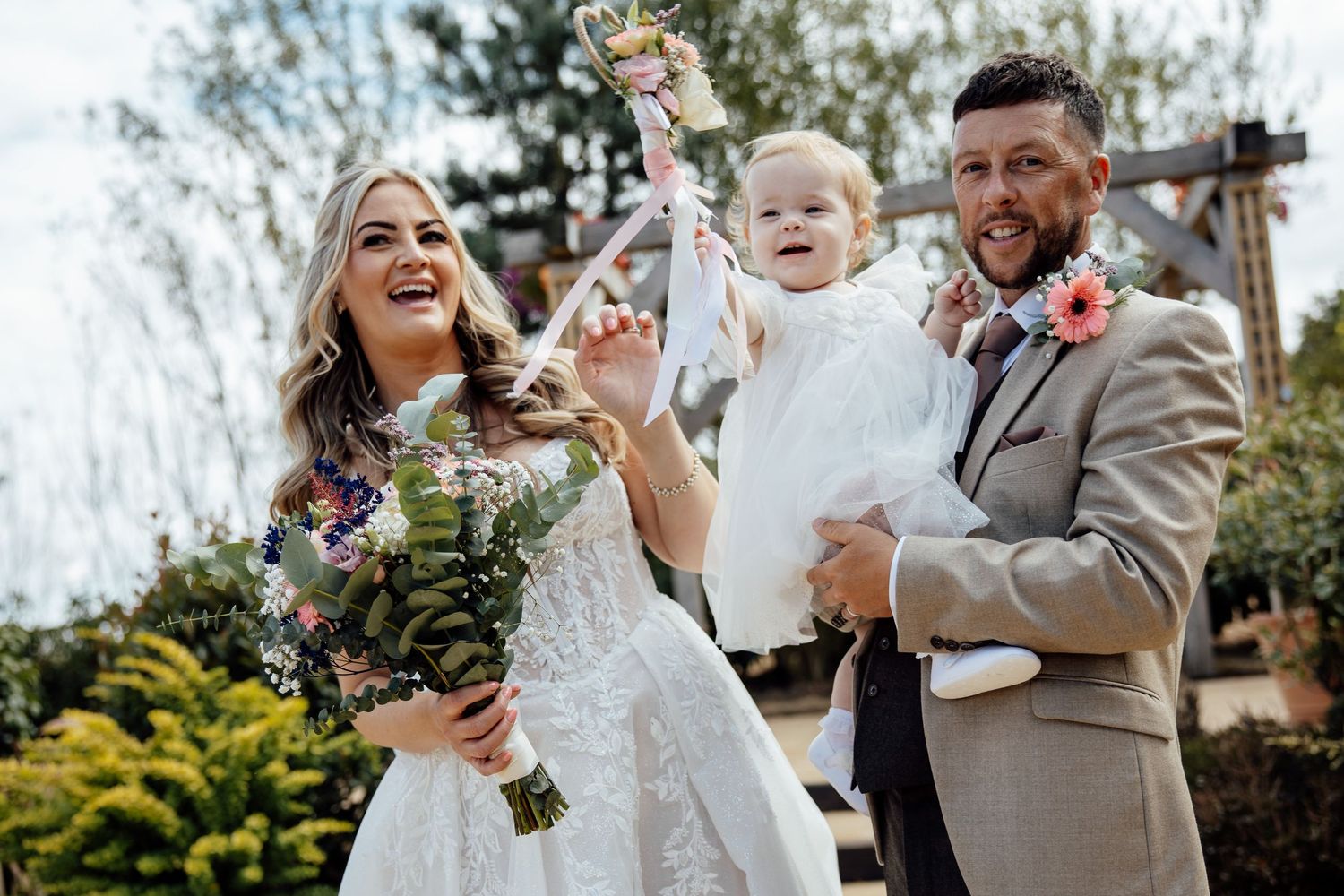 A laughing bride and groom in a tan suit pose with their baby girl holding a floral wand.