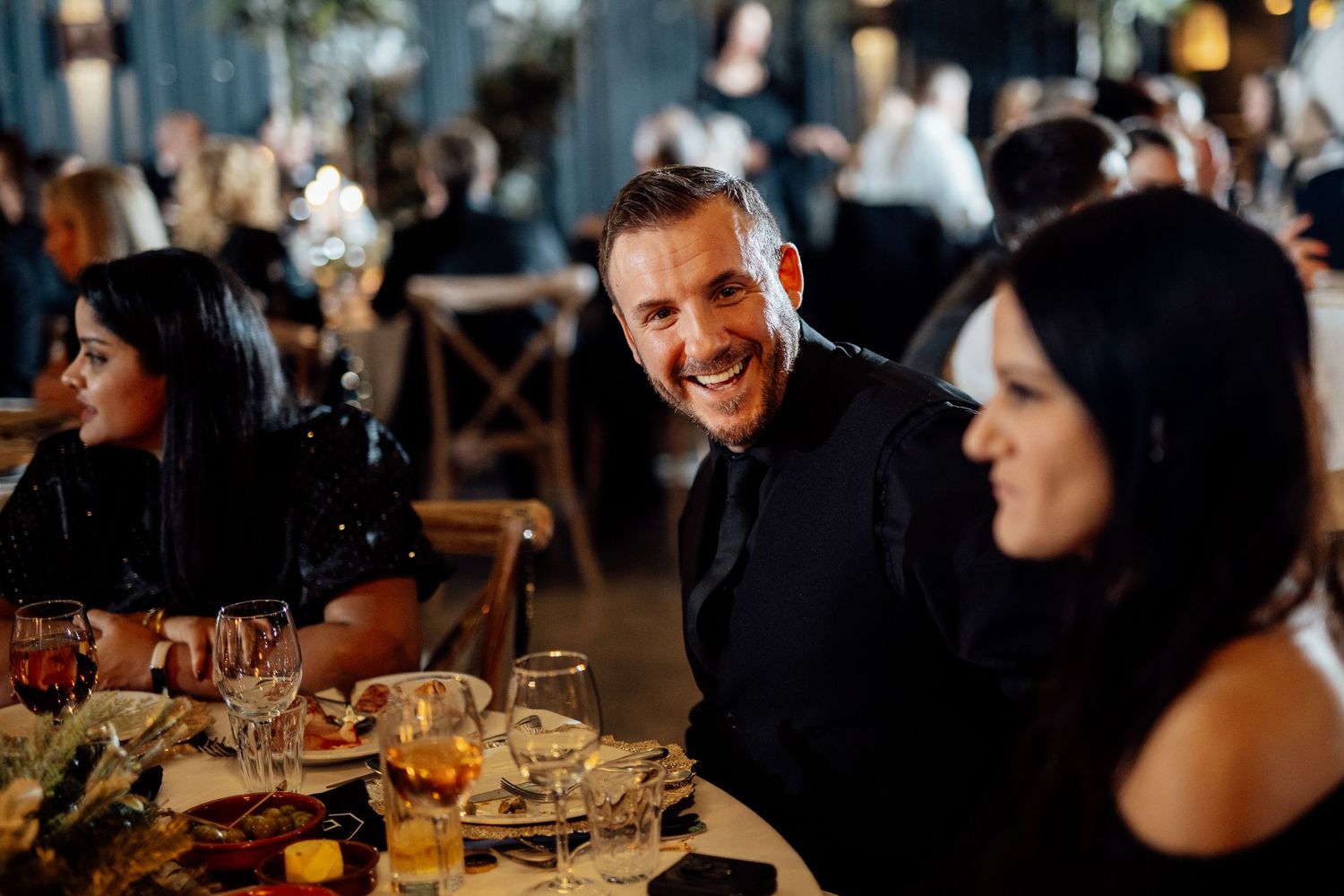Wedding reception guests enjoying dinner and conversation at rustic wooden tables with warm ambient lighting.