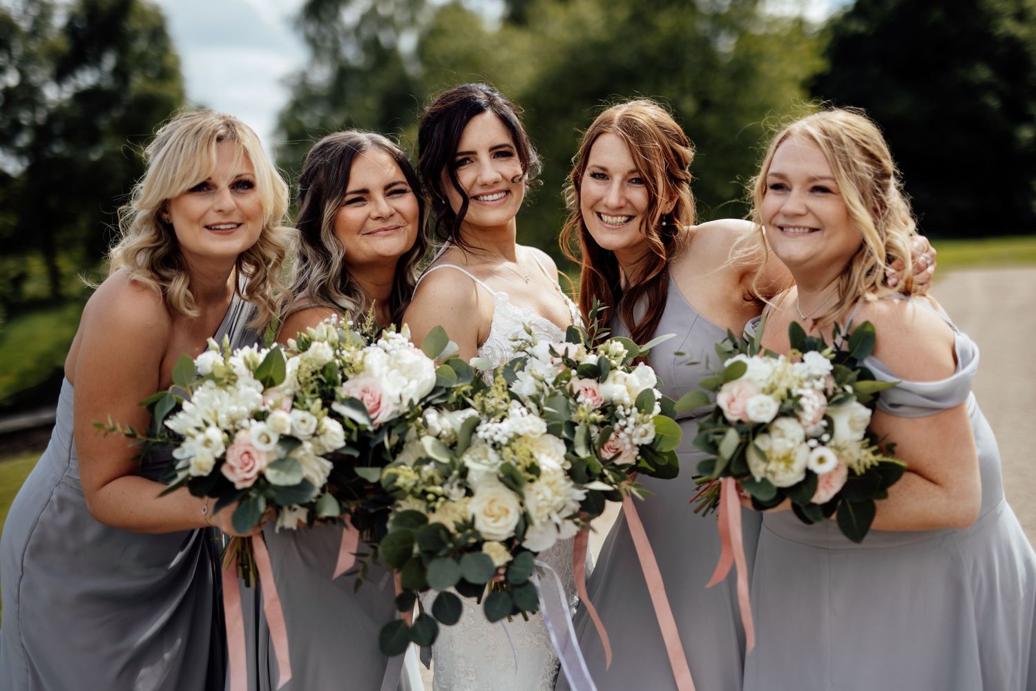 Bridesmaids in gray dresses holding white floral bouquets outdoors.