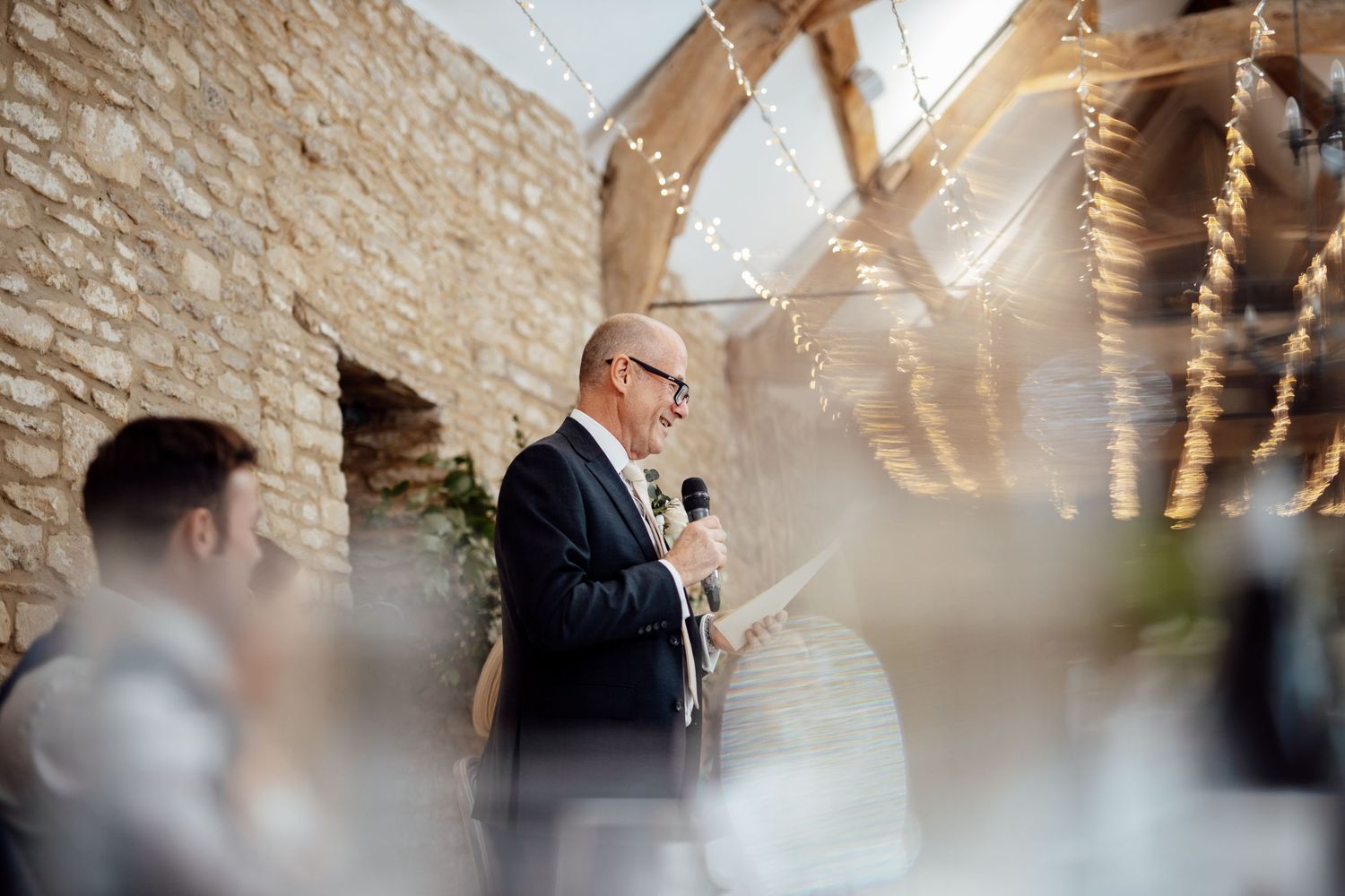 A speaker addresses wedding guests in a stone-walled venue with fairy lights.