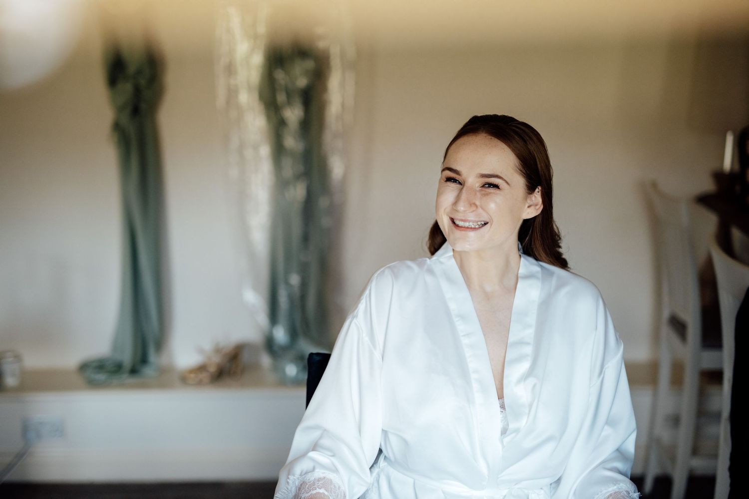 Person in white robe getting ready in dressing room with bridesmaid dresses visible in background.